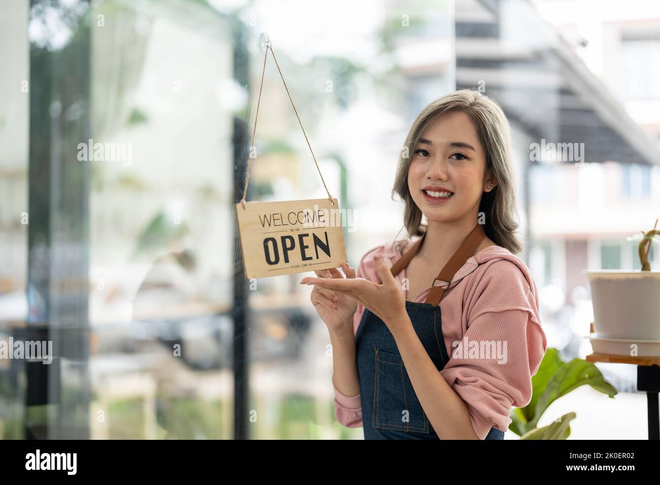 Schöne asiatische junge Barista Frau in Schürze hält Tablet und vor der Tür des Cafés mit offenem Schild stehen. Start des Geschäftsinhabers Stockfoto
