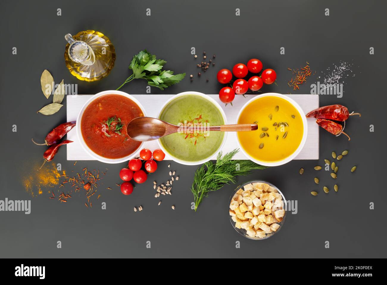 Set aus verschiedenen Gemüsecremesuppen mit Holzlöffel auf einem weißen Holzbrett mit Zutaten auf einem dunkelgrauen Hintergrund. Gemütliches Essen, vegetarische Küche Stockfoto