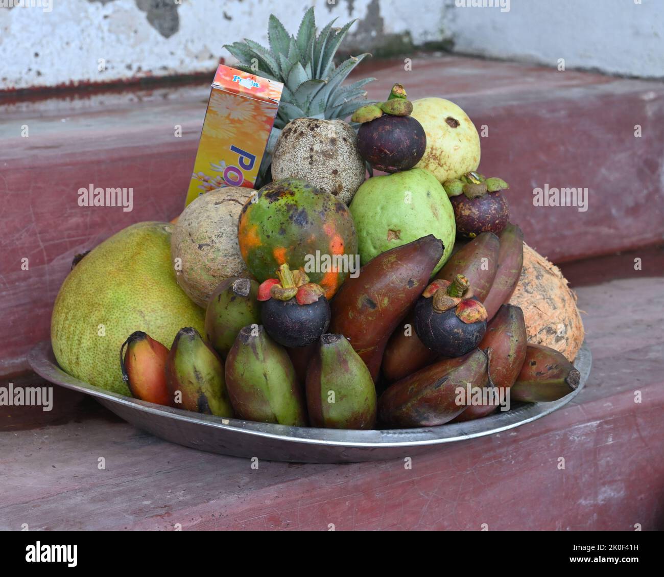 Eine hinduistische Puja-Obstschale voll mit reifen Früchten und einer Schale entfernt und dekoriert Kokosnuss und Räucherstäbchen Paket auf einer kleinen Zementbühne für den Verkauf in der Nähe Stockfoto