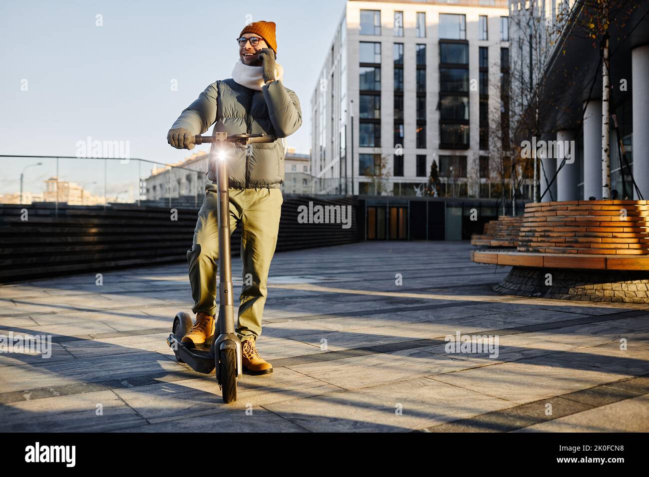 Junger Mann in warmer Kleidung, der während der Fahrt mit dem Motorroller in der Stadt mit dem Handy spricht Stockfoto