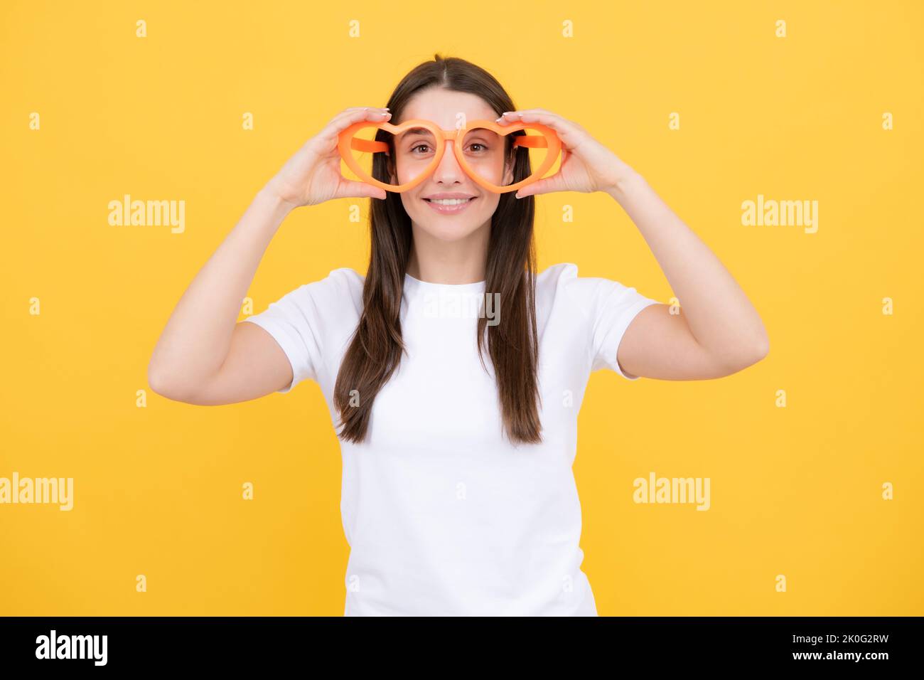 Portrait eines lustigen Mädchens mit coolen Partybrillen. Fröhliches junges Mädchen lächelt mit herzförmigen Gläsern auf gelbem Hintergrund. Stockfoto