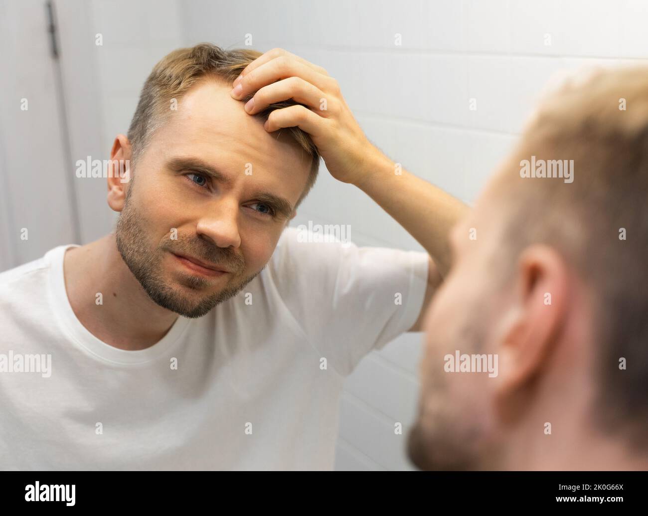 Weißer Mann mittleren Alters mit einem kurzen Bart sieht sich sein Haar im Spiegel im Badezimmer an und macht sich Sorgen wegen einer Glatze. Stockfoto