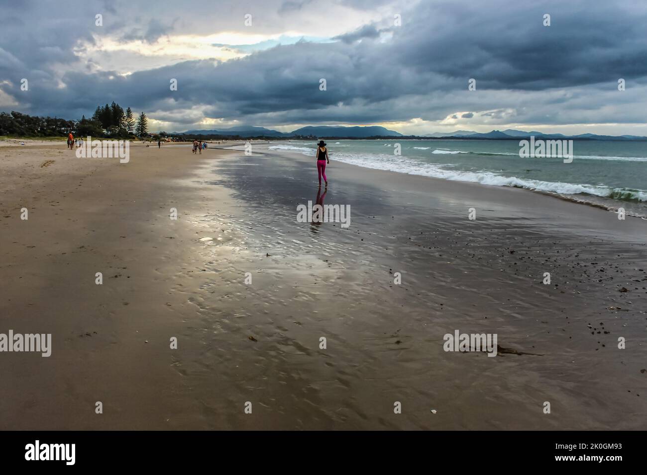 Frau mit rosa Leggings und breitem Hut, die unter stürmischem Himmel am nassen Strand entlang in Byron Bay Australia mit anderen Strandbesuchern und Bergen unterwegs ist Stockfoto