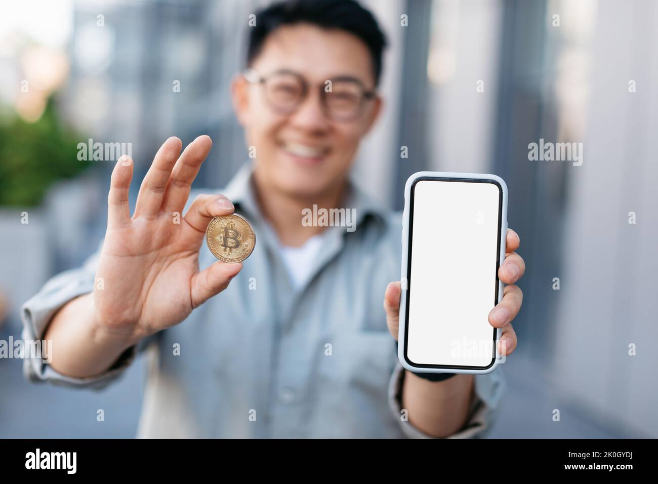 Kryptowährungs-App. Asiatischer Geschäftsmann mit goldener Bitcoin-Münze und leerem Smartphone mit weißem Bildschirm und Mockup Stockfoto