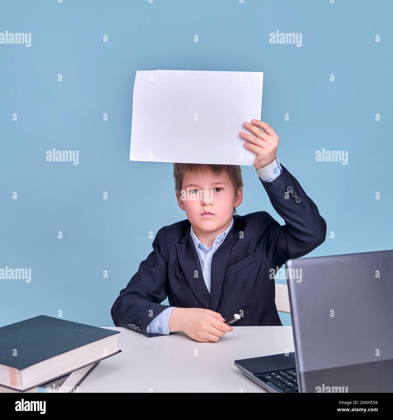 Ein Junge in einem Schulanzug mit einem Blatt A4 Papier an einem Computer während des Fernlernens, kopieren Sie Platz auf einem blauen Studiohintergrund Stockfoto
