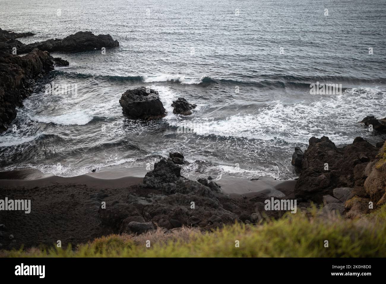 Schwarzer Sandstrand mit vulkanischen Felsen und etwas Gras. Dramatische Landschaft Stockfoto