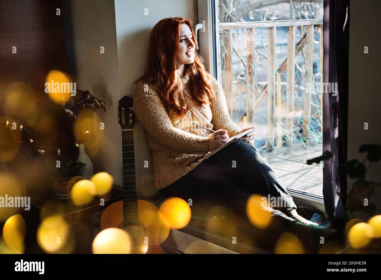 Indoor Cozy Hobbies für Winter, Herbst, kalte Jahreszeit. Redhead Frau spielt akustische Gitarre und hört Musik, sitzt neben Fenster zu Hause Stockfoto