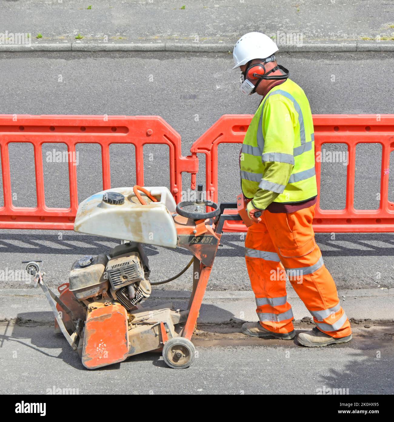 Gut sichtbare Kleidung Gesichtsmaske Ohrenschützer männlichen Arbeiter schieben laut staubigen Scheibe Schneidemaschine bilden Bürgersteig Breitband-Kabel Graben Route Großbritannien Stockfoto