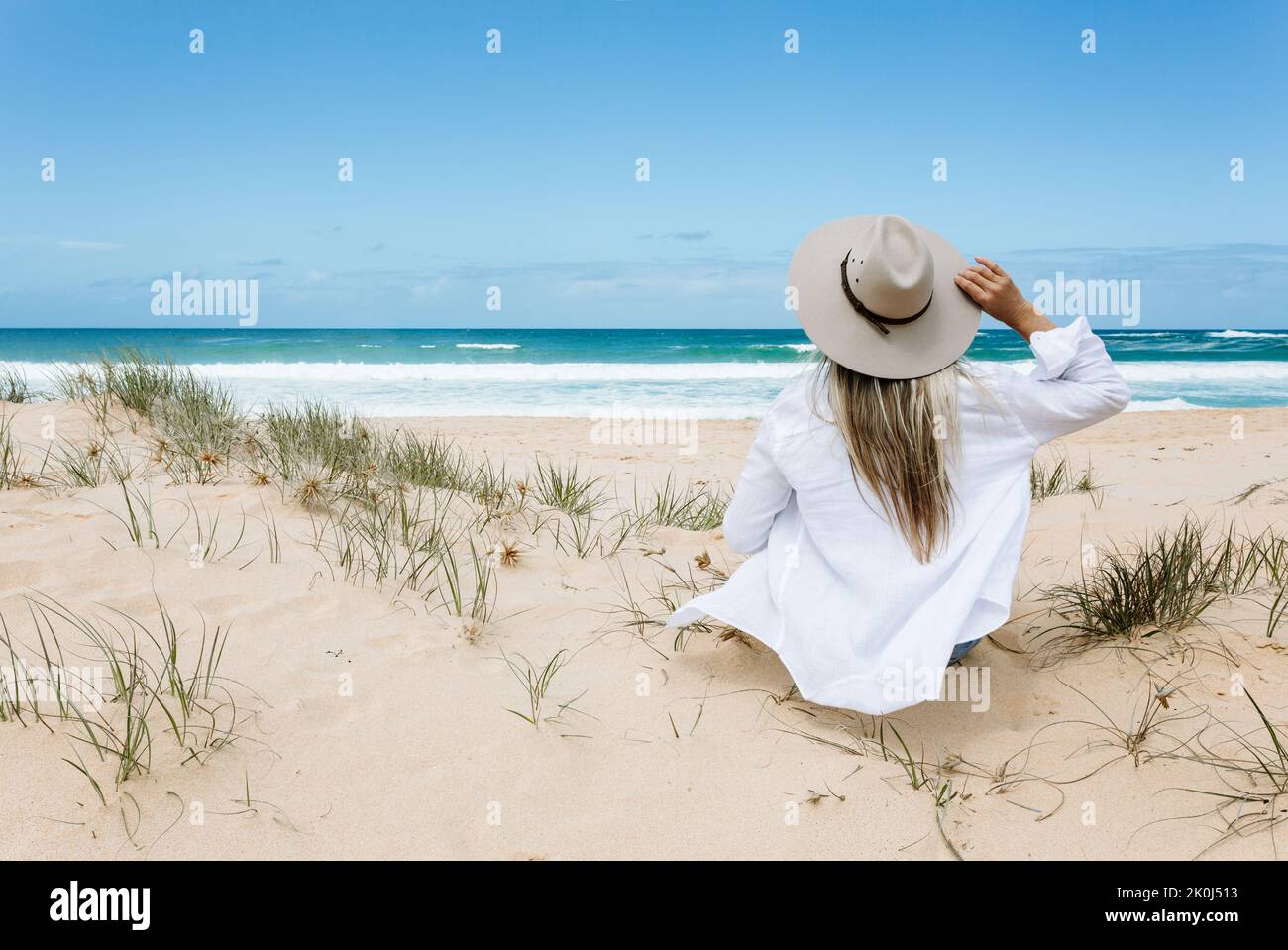 Sorglose Frau im Urlaub im fließenden weißen Hemd über Bikini-Schwimmkostüm, sitzt an einem sauberen Sandstrand mit blauem Himmel. Sie trägt einen Sonnenhut. Stockfoto