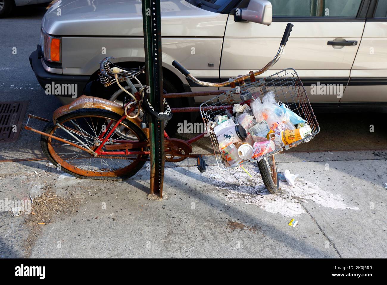 Müll und Abfall im Lenkerkorb eines rostigen verlassenen Fahrrads, das in New York City an eine Stange gekettet ist. Stockfoto