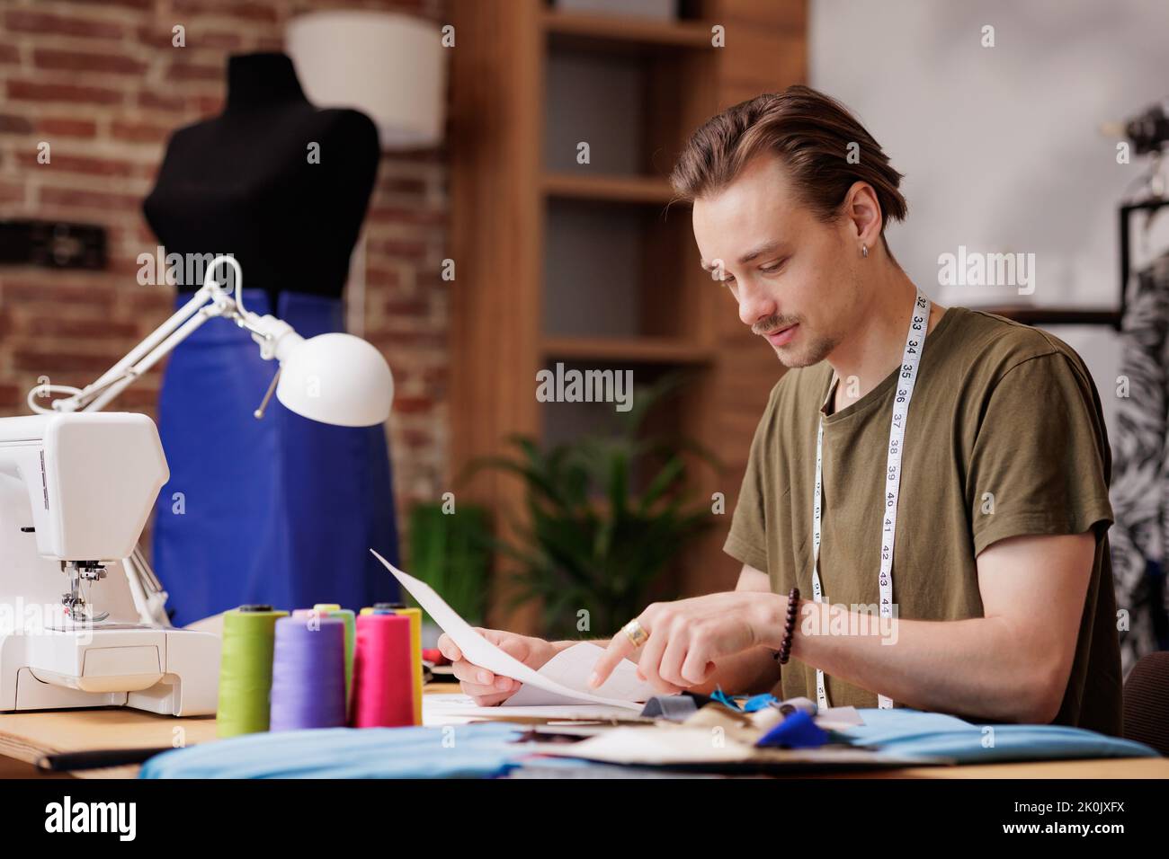 Ein junger Mann ist ein Modedesigner. Er schaut sich die Skizze an, während er mit einer Nähmaschine an einem Tisch sitzt Stockfoto