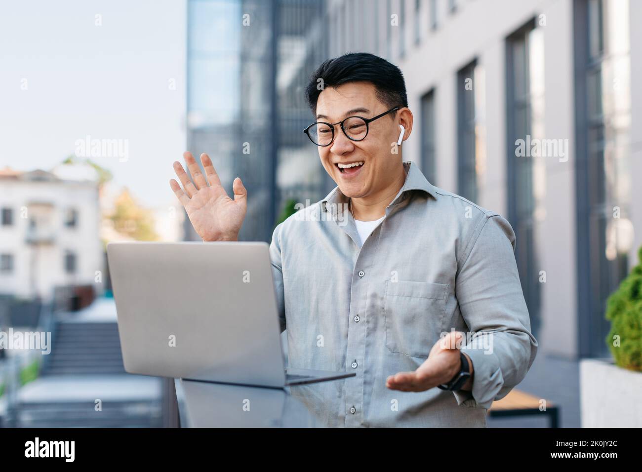 Aufgeregt asiatischen Geschäftsmann mit Videoanruf auf Laptop, im Gespräch mit Webcam, sitzen im Freien in der Nähe von modernen Bürogebäude Stockfoto