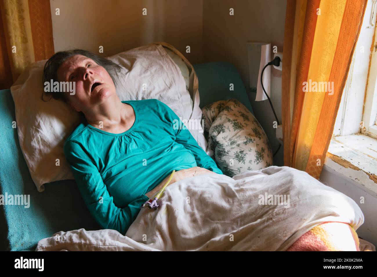 Ältere Frau mit Lähmung, die unter der Decke auf einem medizinischen Bett in der Nähe der Fenster in einem schäbigen Zimmer zu Hause lag Stockfoto