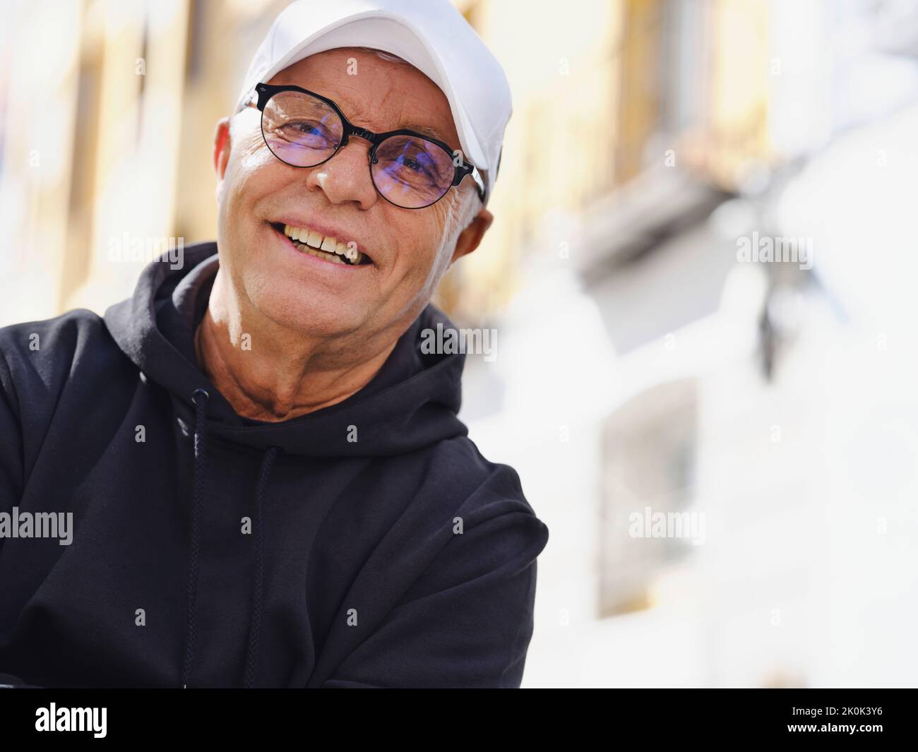 Hübscher älterer Mann in trendigen Brillen und stilvoller Kleidung, der auf unscharfen Hintergrund der Straße die Kamera anschaut Stockfoto