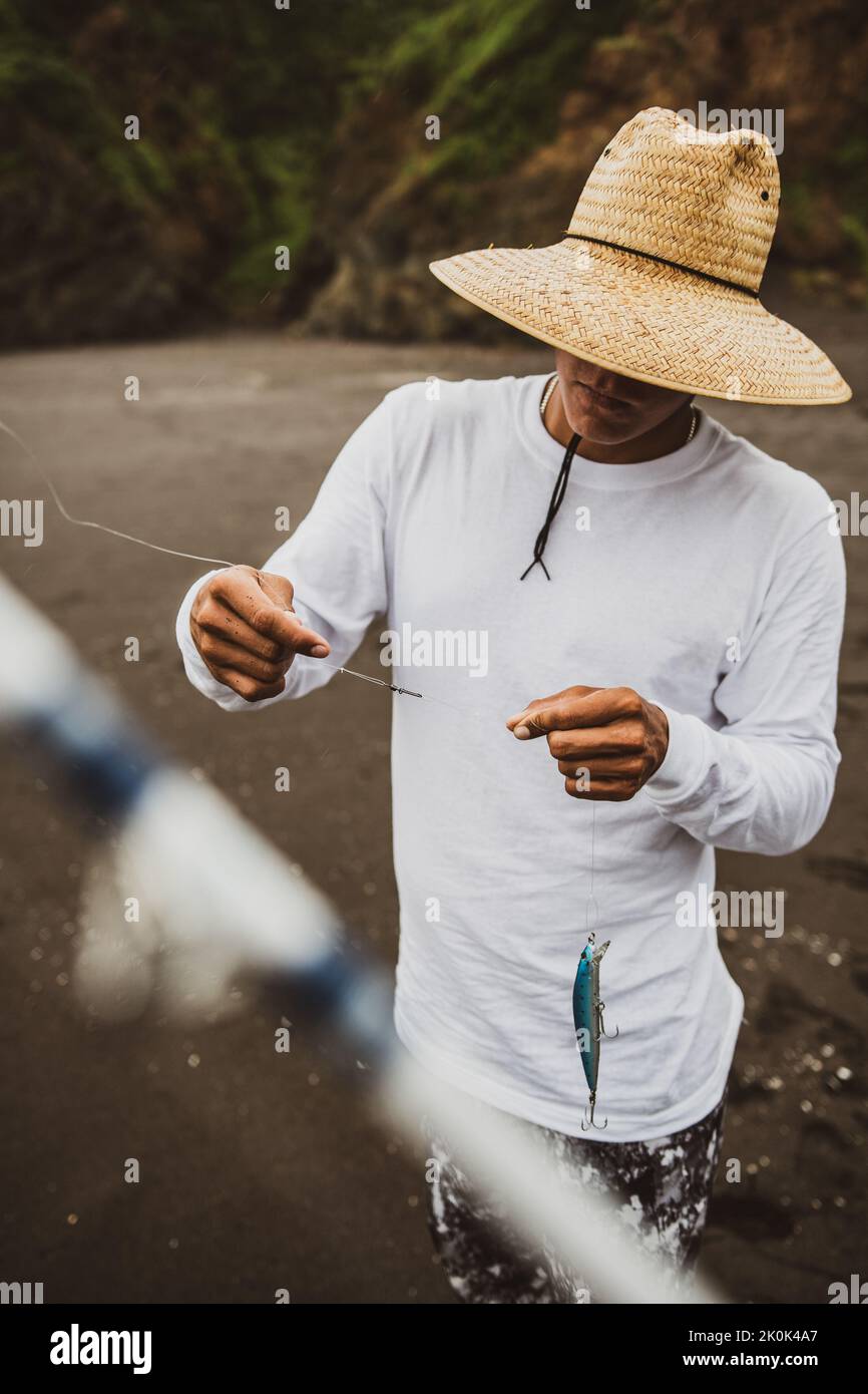 Nicht erkennbarer Rüde im Hut, der einen speziellen Stecker an der Angelschnur bindet, während er an der Küste mit Angelrute im Küstengebiet steht Stockfoto