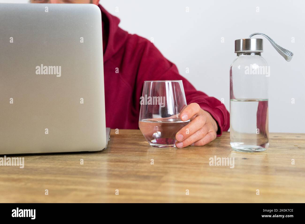 Ein nicht erkennbarer männlicher Arbeiter, der im Netbook im Internet surft, während er während der Arbeit im hellen Büro mit einer Flasche Wasser am Schreibtisch sitzt Stockfoto