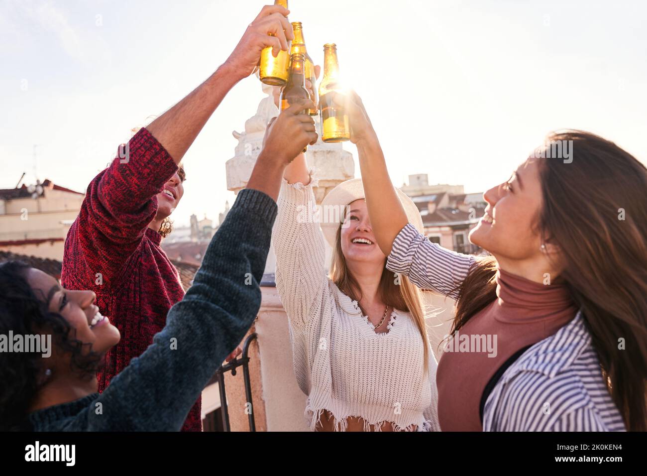Gruppe von zufriedenen jungen Freunden klirren Flaschen mit kaltem Bier, während auf der sonnigen Terrasse Stockfoto