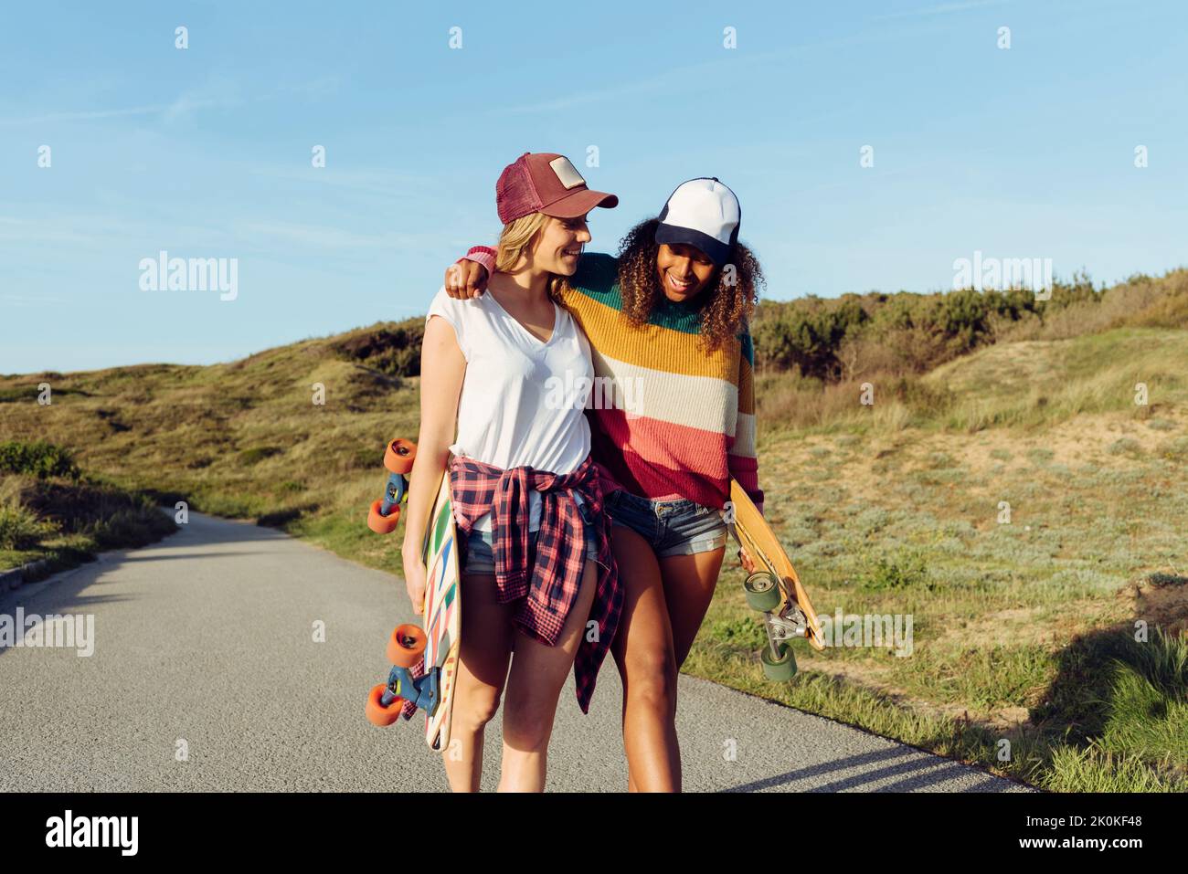 Zwei wunderschöne, multiethnische, fröhliche Frauen, die Skateboards halten, während sie an einem sonnigen Tag mitten auf einer Straße gehen und sich auf das Sho des anderen lehnen Stockfoto