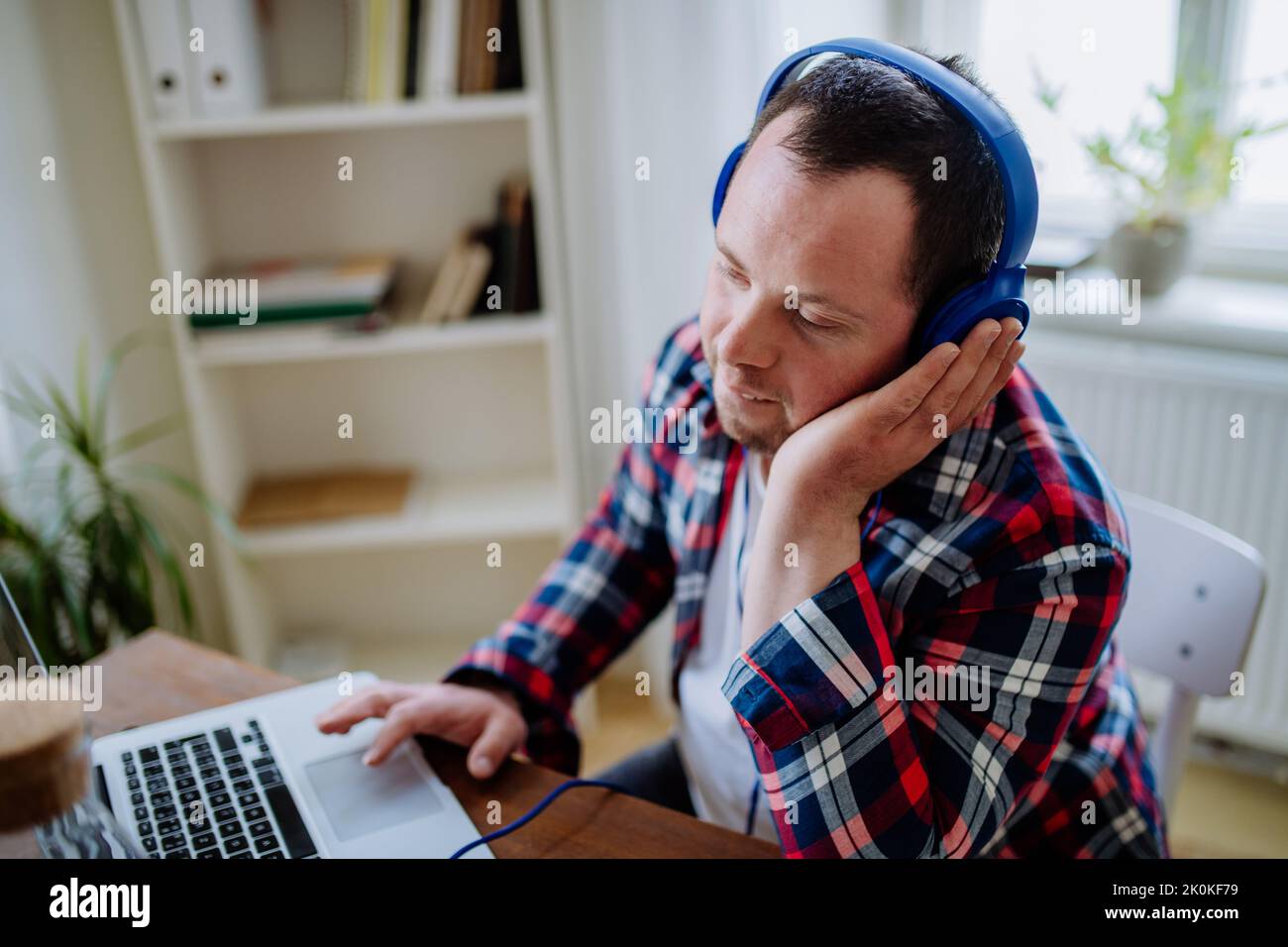 Junger Mann mit Down-Syndrom, der im Büro am Schreibtisch sitzt und mit einem Laptop Musik von Kopfhörern hört. Stockfoto