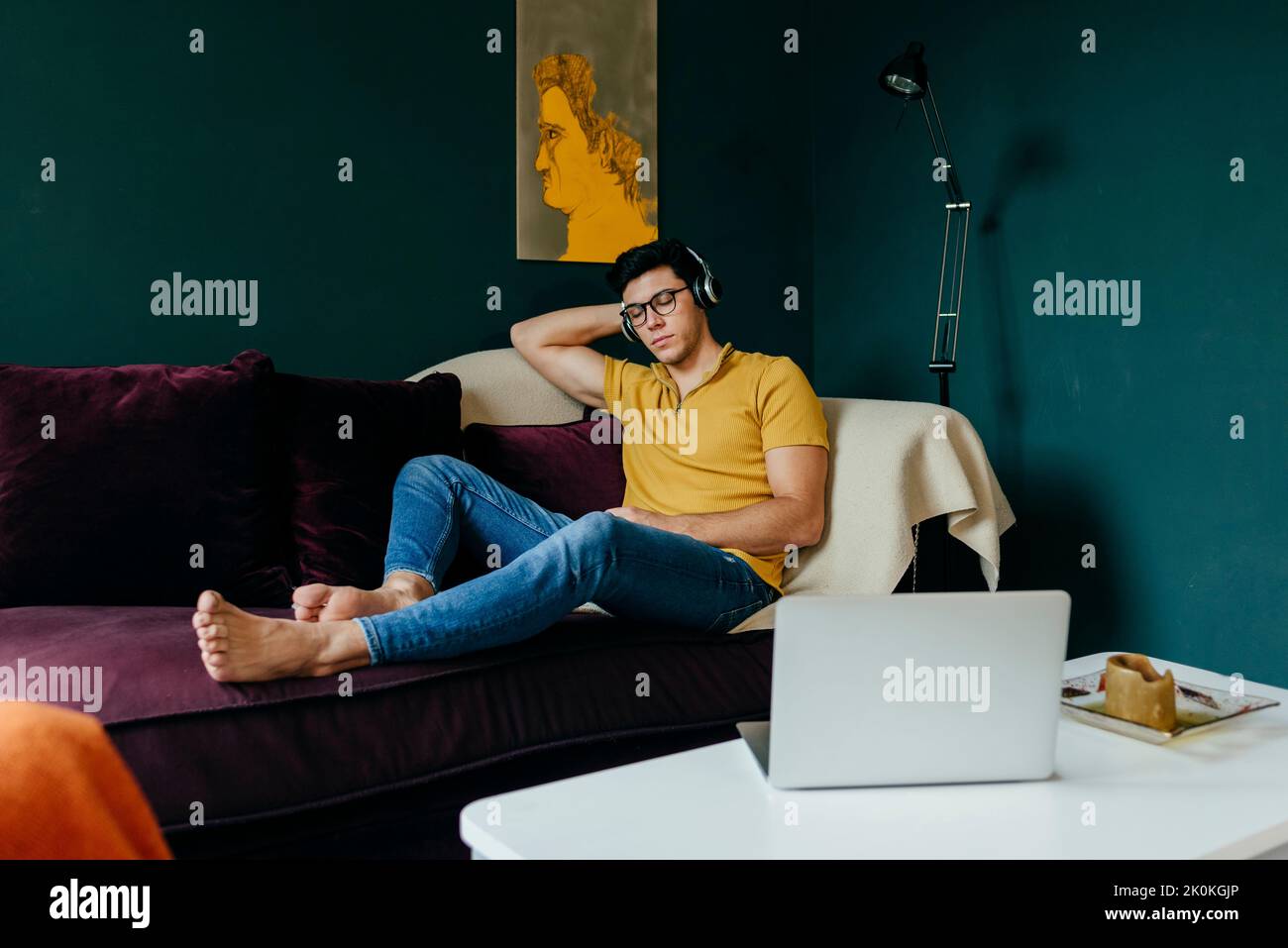 Attraktiver kaukasischer Mann, der mit geschlossenen Augen auf der Couch sitzt, während er zu Hause ein Nickerchen mit Musik über Kopfhörer macht Stockfoto