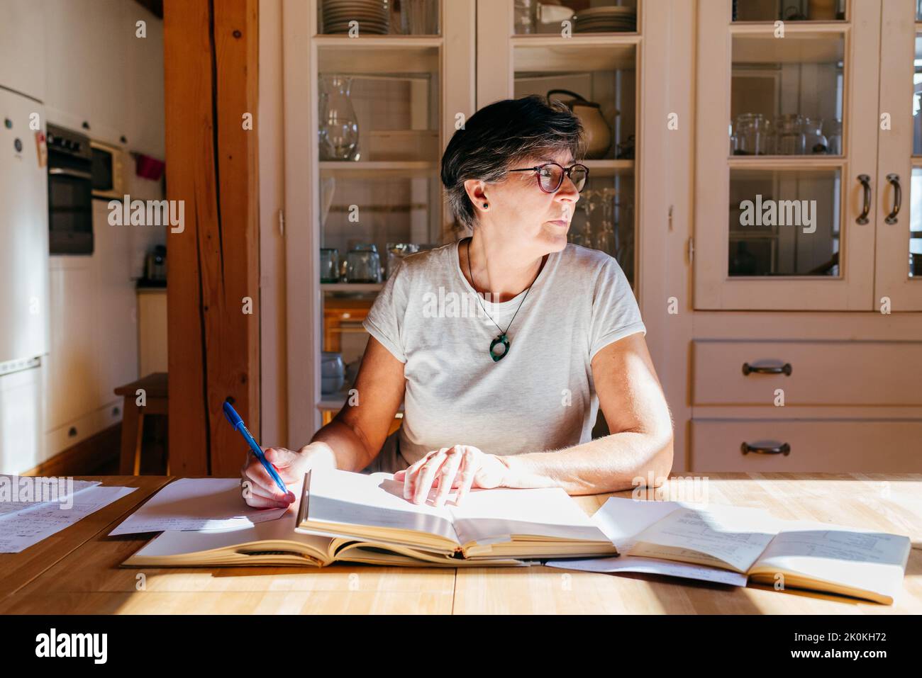 Fokussierte Frau mittleren Alters in einer Brille mit Stift in der Hand, die mit einem Haufen Bücher und Papiere am Tisch sitzt und zu Hause professionelle Materialien studiert Stockfoto