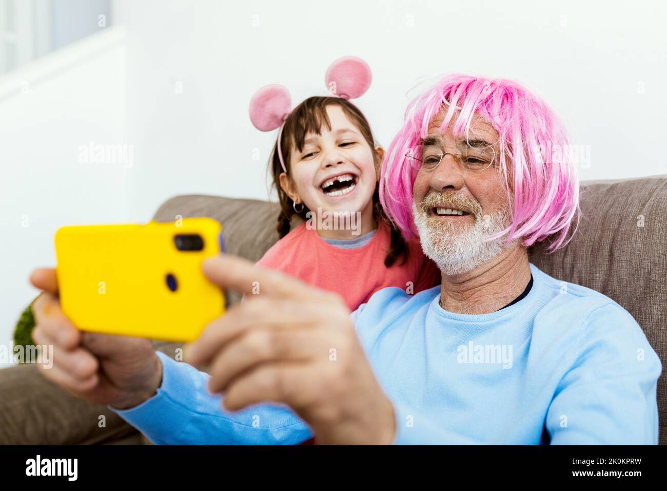 Glückliches Kind, das auf der Couch mit Großvater mit rosa Perücke mit falschen Ohren sitzt und Selfie auf dem Smartphone im Zimmer macht Stockfoto