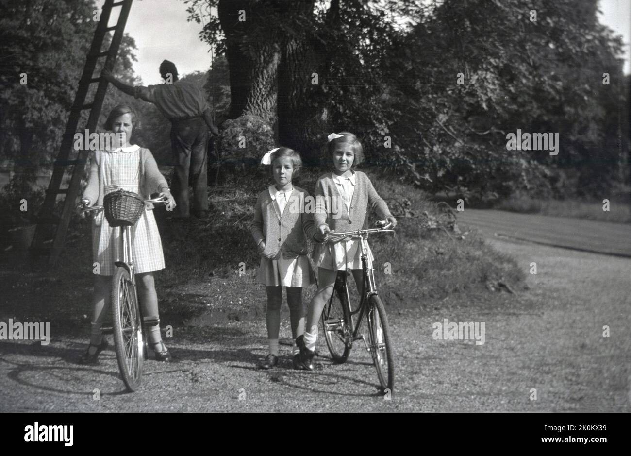1937, historisch, drei Schulmädchen, zwei auf Fahrrädern, auf einem Schotterweg auf dem Gelände des Landgutes Forcett Hall, North Yorkshire, England, Großbritannien. Im Hintergrund befindet sich ein Gutsgärtner mit einer Leiter bei einem Baum. Stockfoto
