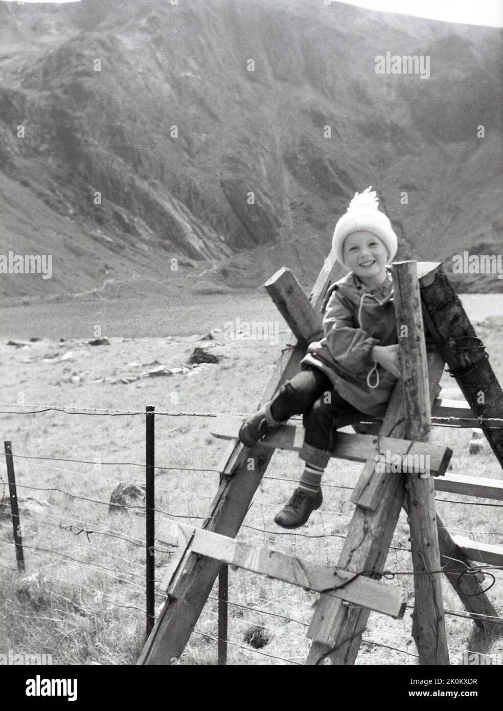 1956, historisch, ein Kind sitzt für ein Foto auf einem Holzstiel in einem Feld, Berge im Hintergrund, Snowdonia, Wales, Großbritannien. Stile sind Strukturen, die mit Stufen oder Leitern in ländlichen Gebieten hergestellt werden, die Spaziergängern ermöglichen, über einen Zaun zu gehen, während sie Tiere schützen und ihr Entweichen verhindern. Stockfoto