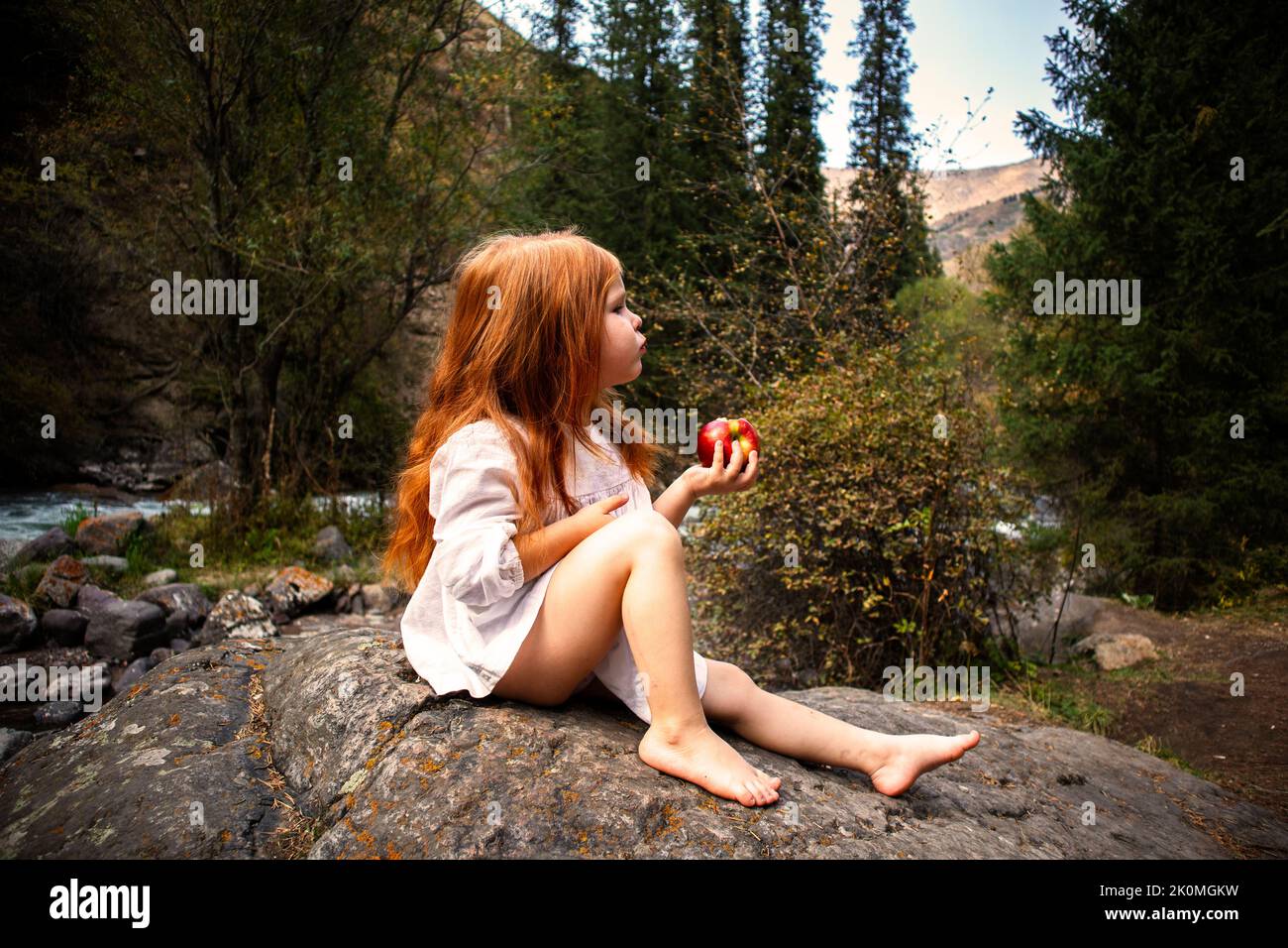 Ein kleines Mädchen mit langen roten Haaren in einem weißen Kleid sitzt am Fluss und isst einen Apfel Stockfoto