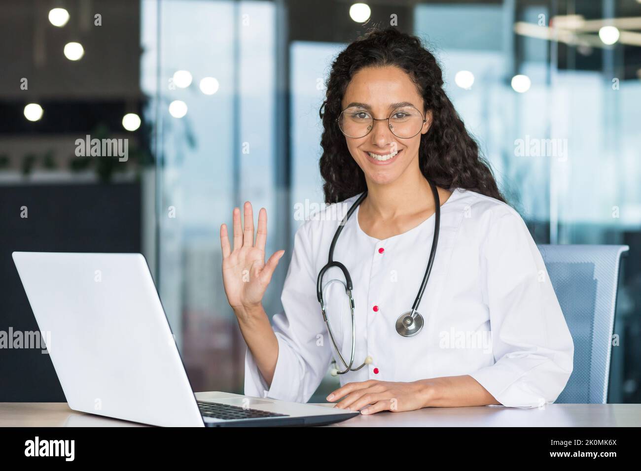 Porträt einer jungen professionellen lateinamerikanischen Ärztin in einem weißen medizinischen Mantel lächelnd und in die Kamera schauend, hält die Ärztin ihre Hand hoch und grüßt sie Stockfoto