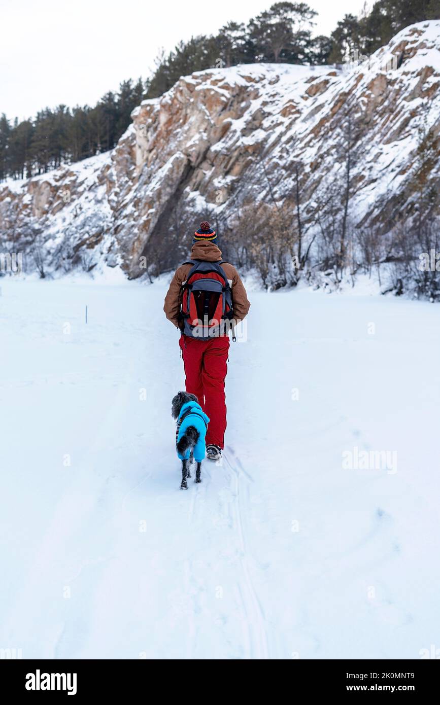 Rückansicht junger Mann in rotbrauner warmer Kleidung mit Rucksack mit Mischlingshund in warmem blauem Anzug, der im Winter AC zwischen Felsen und Klippen im Schnee läuft Stockfoto