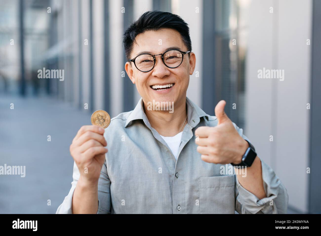 Ein glücklicher asiatischer Geschäftsmann, der im Freien in der Nähe des modernen Bürogebäudes Bitcoin zeigt und den Daumen hochgestikelt Stockfoto
