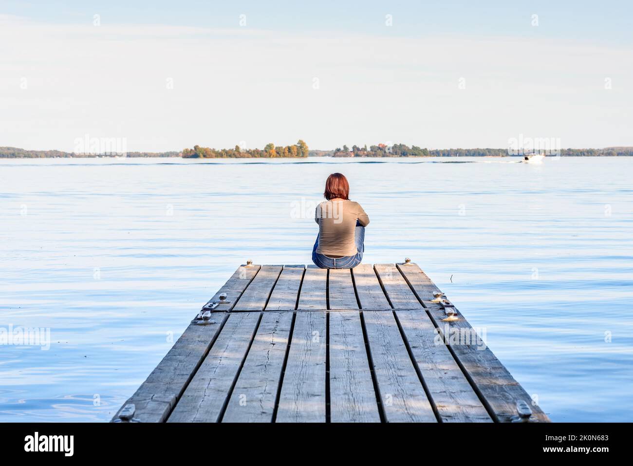 Einsame Frau, die an einem sonnigen Herbsttag am Ende eines hölzernen Steges am Ufer eines großen Flusses sitzt. Konzept der Einsamkeit. Stockfoto