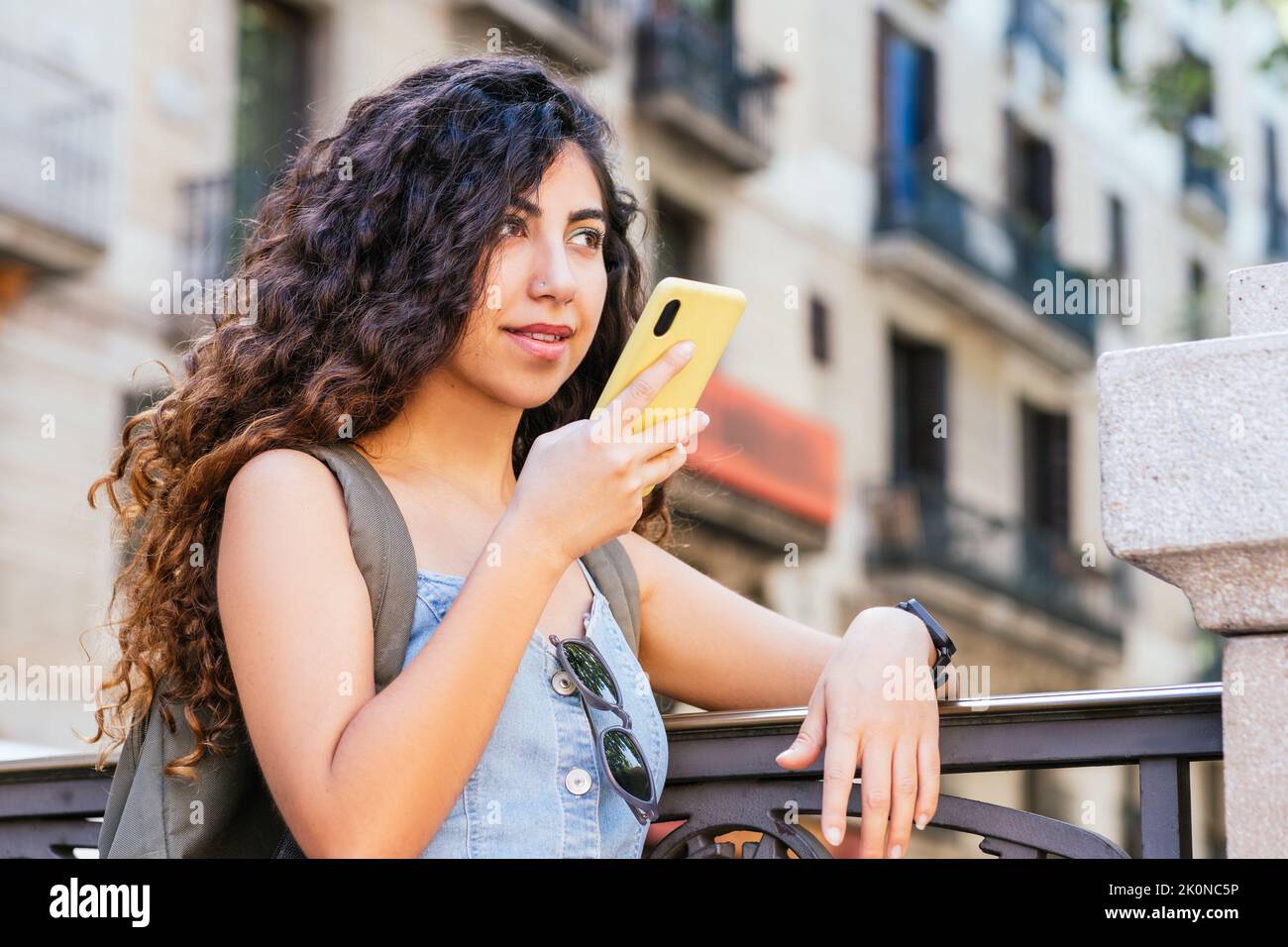 Indische junge Frau, die sich an einen Zaun lehnt und mit ihrem gelben Smartphone eine Sprachnachricht sendet Stockfoto