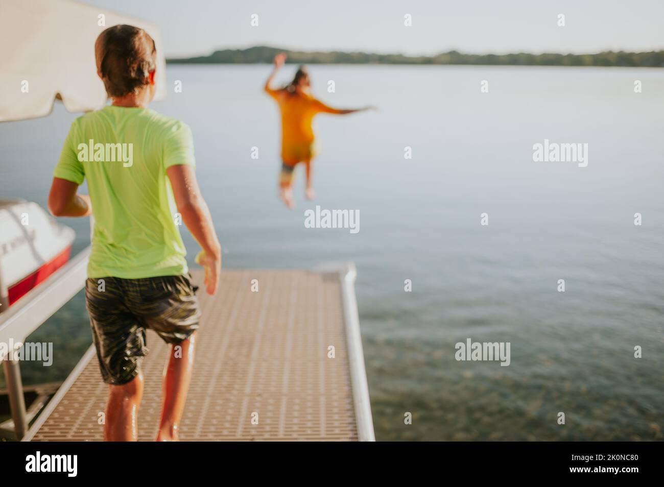 Ein Kind springt von einem Dock ins Wasser, während das andere vom Dock herunterläuft Stockfoto