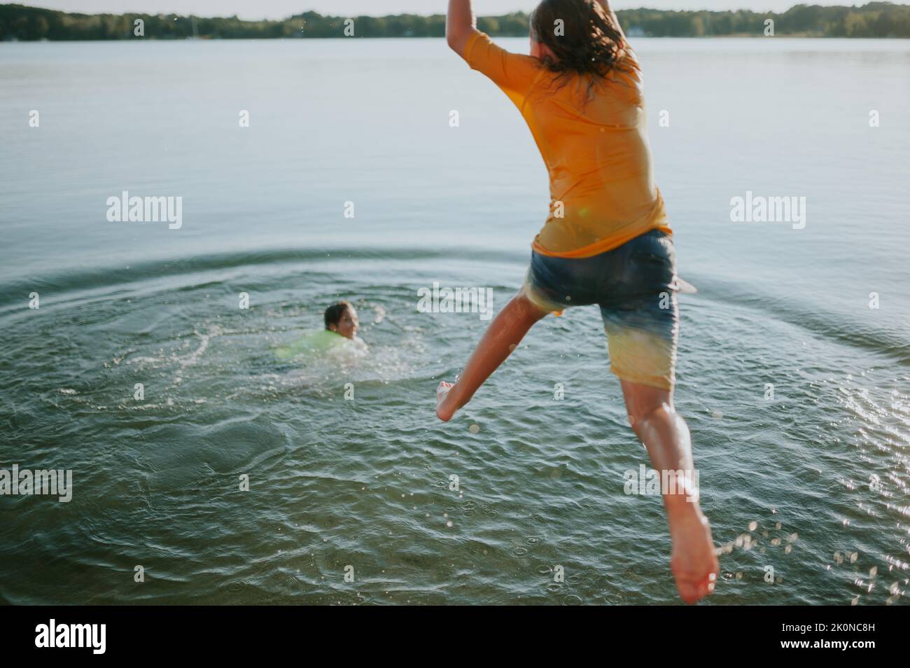 Junge springt vom Dock, während ein anderer Junge lächelt und im Wasser schwimmend Stockfoto