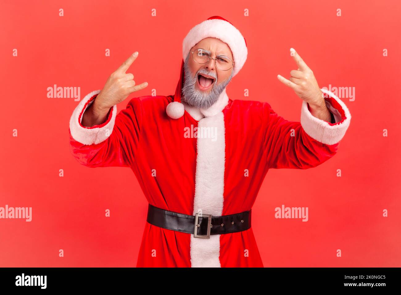 Aufgeregt älterer Mann mit grauem Bart trägt weihnachtsmann Kostüm schreiend und zeigt Rock and Roll Zeichen, Blick auf die Kamera, Weihnachten zu feiern. Innenaufnahme des Studios isoliert auf rotem Hintergrund. Stockfoto