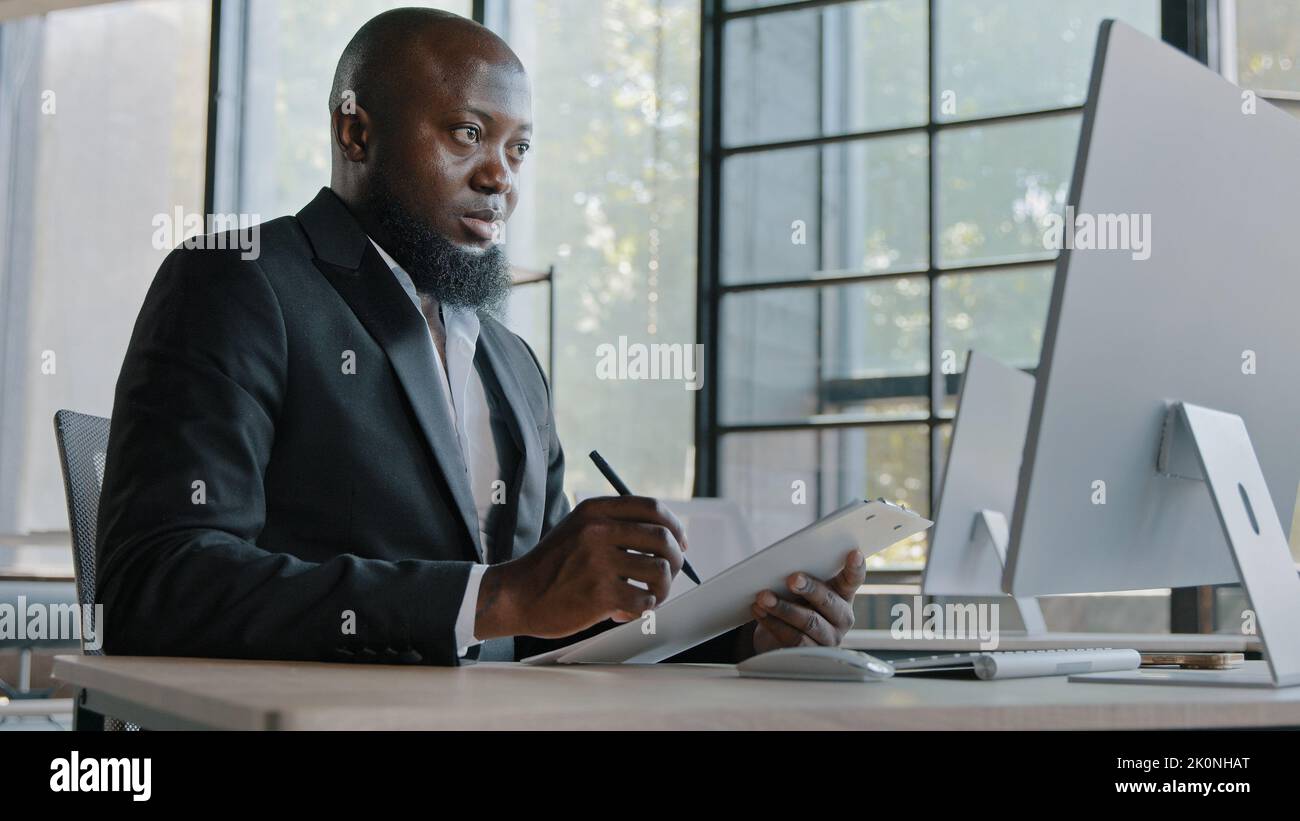 Senior Geschäftsmann afroamerikanischen Rechtshänder Mitarbeiter Designer sitzen im Büro Blick auf Computer-Bildschirm schreiben neue Informationen verwenden Papier Tablet Stockfoto