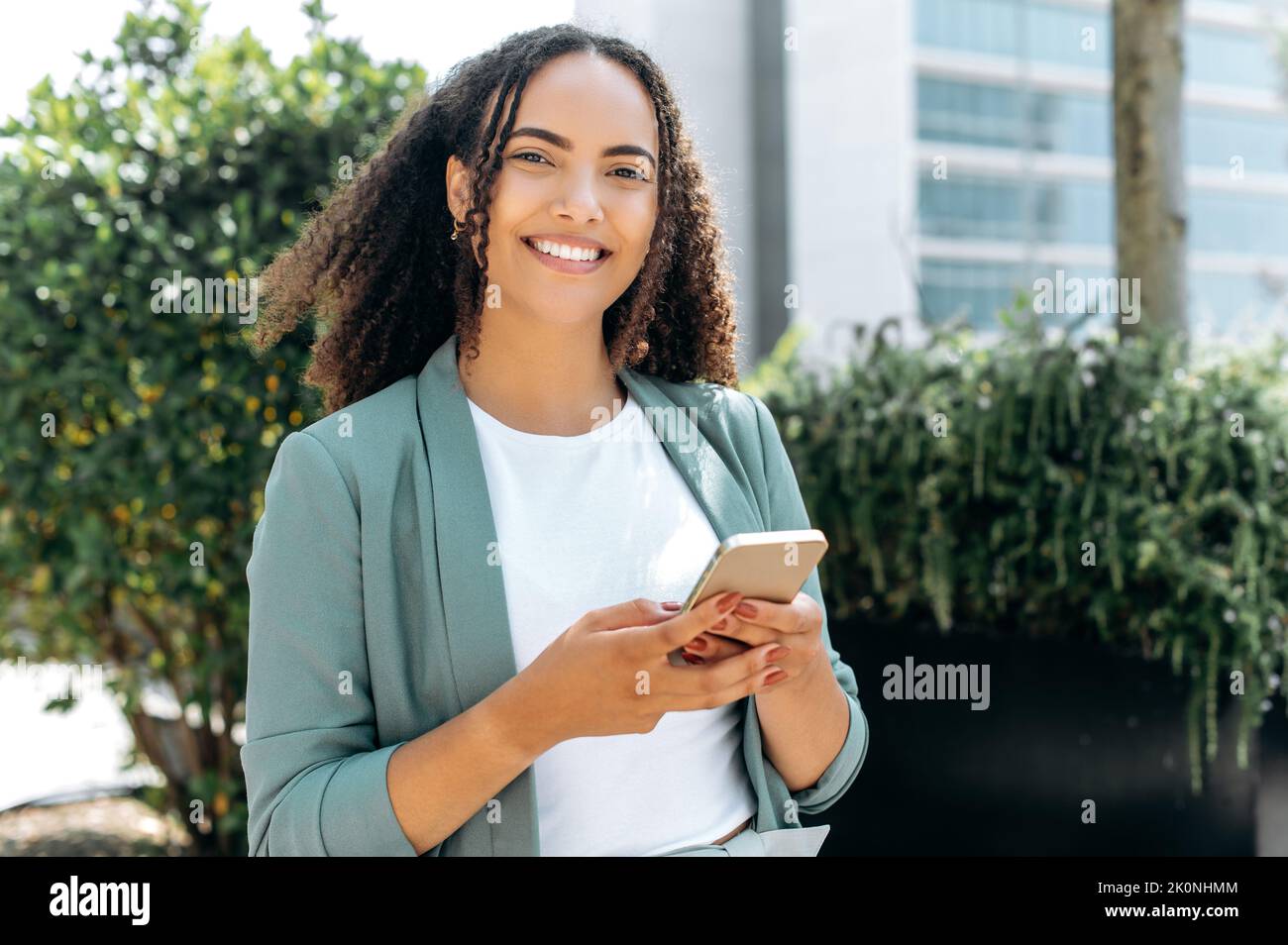 Gadget wird verwendet. Positive hispanische oder brasilianische lockige junge Frau, stilvoll gekleidet, mit ihrem Handy, steht im Freien, Messaging online mit Freunden in den sozialen Medien, schaut auf die Kamera, lächelnd Stockfoto