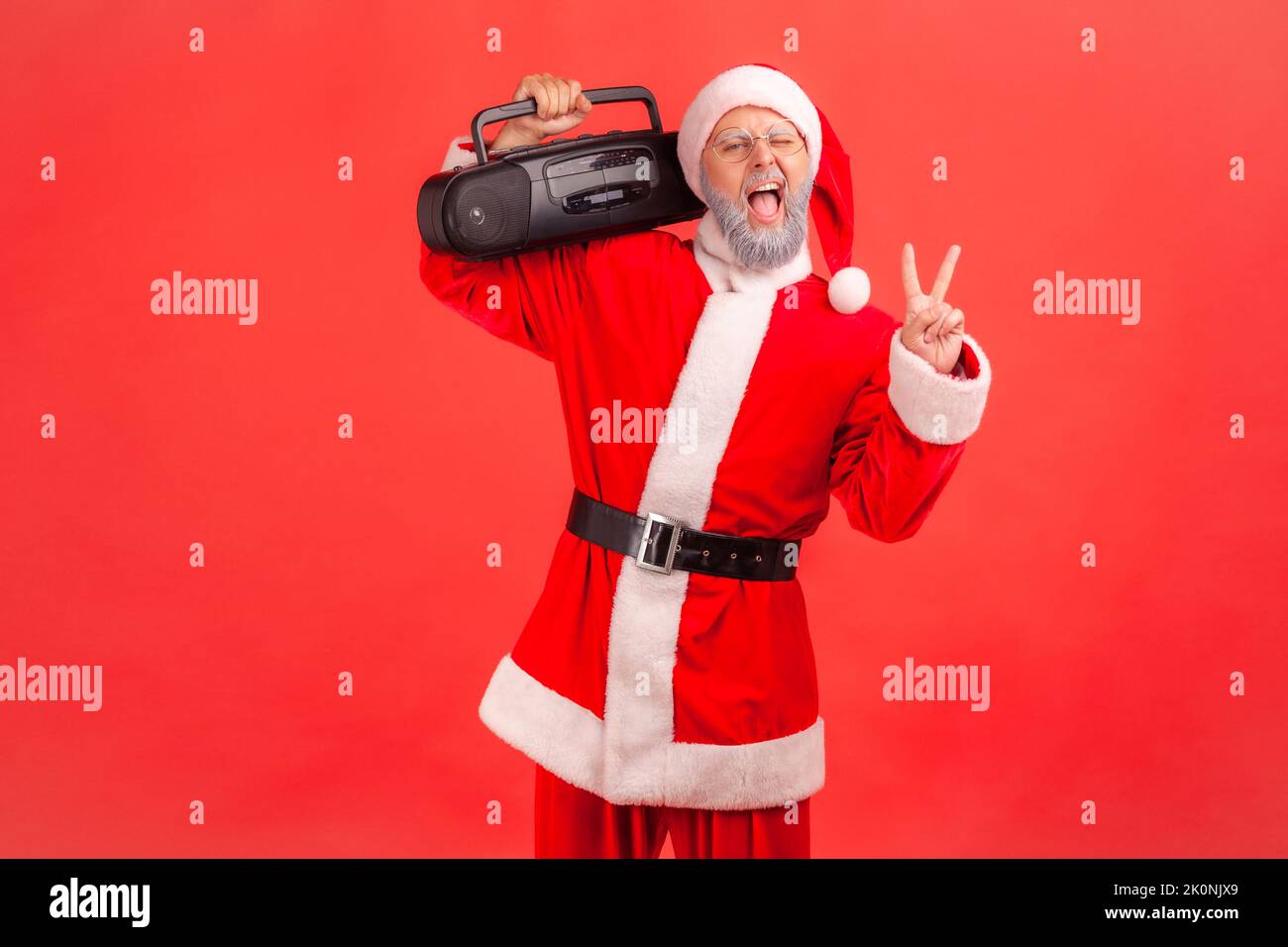 Positive lustige ältere Mann mit grauem Bart trägt weihnachtsmann Kostüm stehend und hält Tonbandgerät zeigt V-Zeichen, feiert Weihnachtsfeier. Innenaufnahme des Studios isoliert auf rotem Hintergrund. Stockfoto