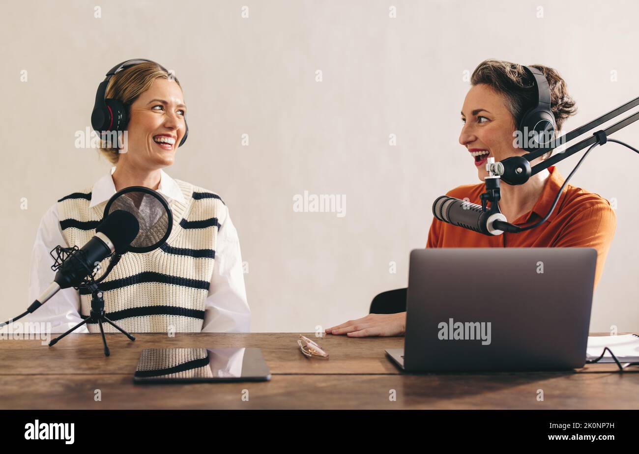 Glückliche weibliche Podcasterinnen, die bei einer Audioübertragung in einem Heimstudio ein tolles Gespräch führen. Zwei fröhliche Frauen, die einen Internet-Podcast für ihre Aufnahmen Stockfoto