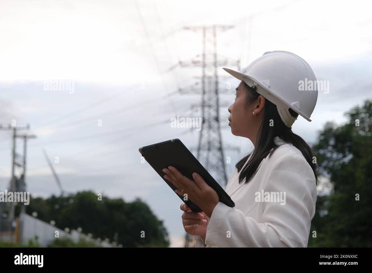 Asiatische Elektroingenieurin, die an einem Tablet in der Nähe des Hochspannungsmastes arbeitet und das Stromnetz inspiziert. Windenergieanlageningenieur für die Stromerzeugung. Stockfoto