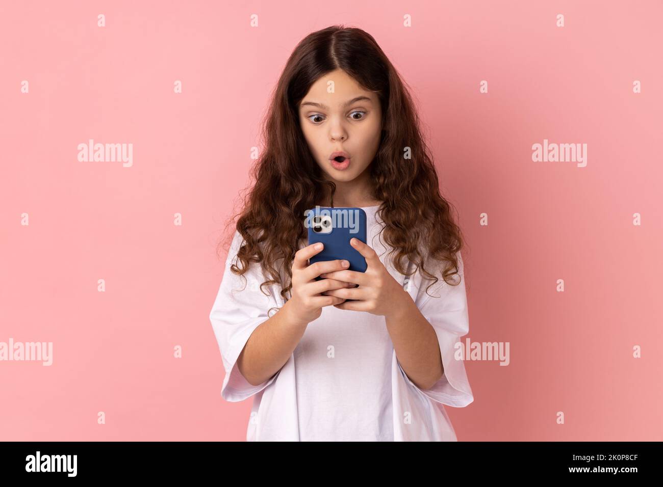 Mobile Verbindung. Porträt eines schockierten erstaunten kleinen Mädchens mit weißem T-Shirt und einem schockierenden Beitrag über das Handy in einem sozialen Netzwerk. Innenaufnahme des Studios isoliert auf rosa Hintergrund. Stockfoto
