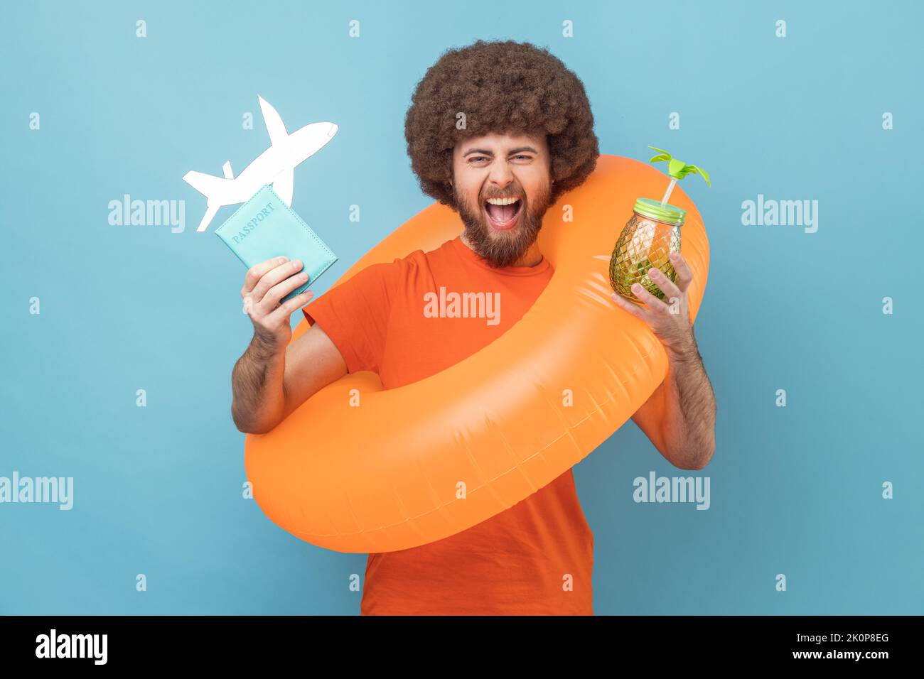 Mann mit Afro-Frisur, der mit orangefarbenem Gummiring steht, Passdokument und Flugzeugmodell hält, Cocktail trinkt und Reisetour freut. Innenaufnahme des Studios isoliert auf blauem Hintergrund. Stockfoto