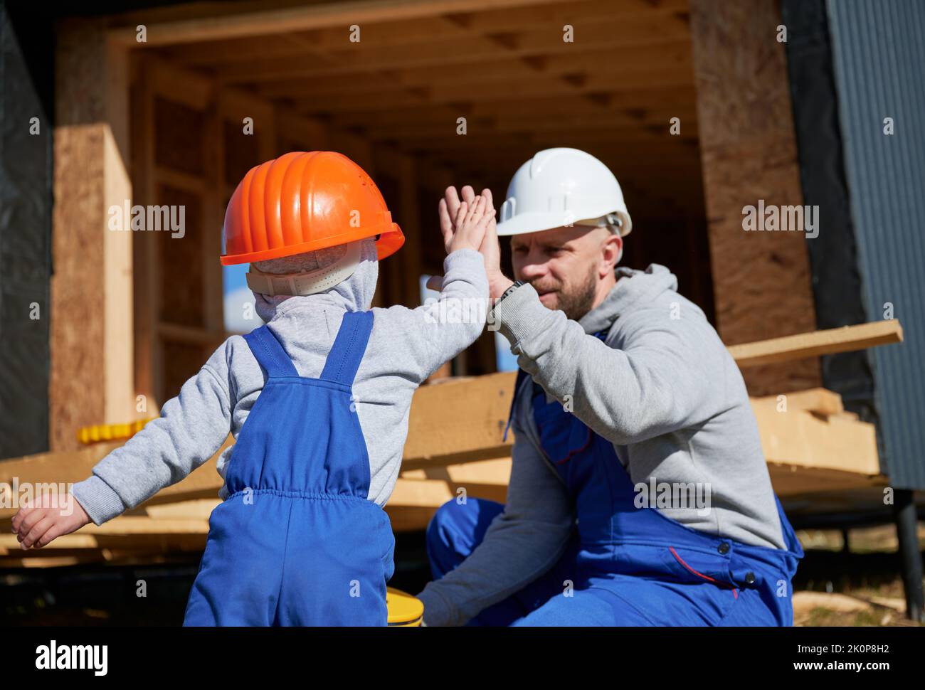 Vater mit Kleinkind Sohn Gebäude Holzrahmen Haus. Der männliche Baumeister gab dem Kind auf der Baustelle hohe fünf und trug am sonnigen Tag Helm und blaue Overalls. Zimmerei- und Familienkonzept. Stockfoto