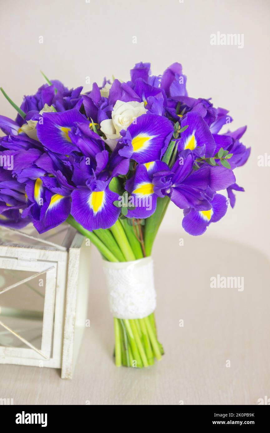 Blumenstrauß aus Iris mit weißen Rosen und weißer dekorativer Laterne auf beigem Hintergrund. Nahaufnahme, Vorderansicht, vertikales Foto. Stockfoto