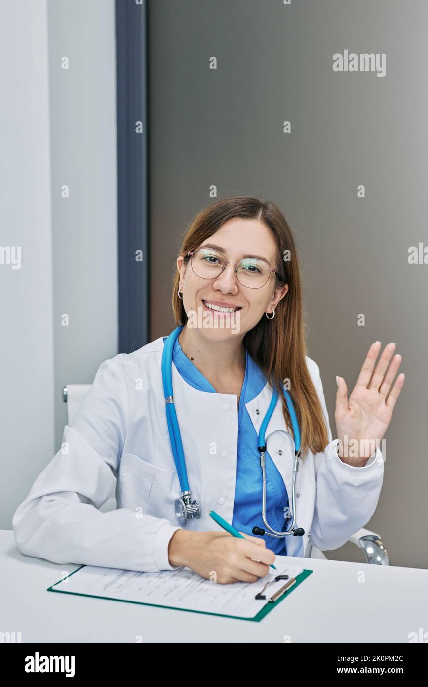 Freundliche Ärztin, die in medizinischer Uniform an seinem Arbeitsplatz sitzt und im Krankenhaus mit der Hand winkt Stockfoto