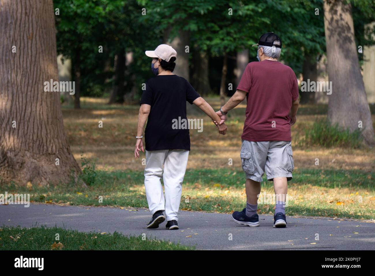 Ein älteres Paar, das im Park spazieren ging, wobei der Mann die Frau für zusätzliche Unterstützung und Stabilität festhielt. In Flushing, Queens, New York. Stockfoto