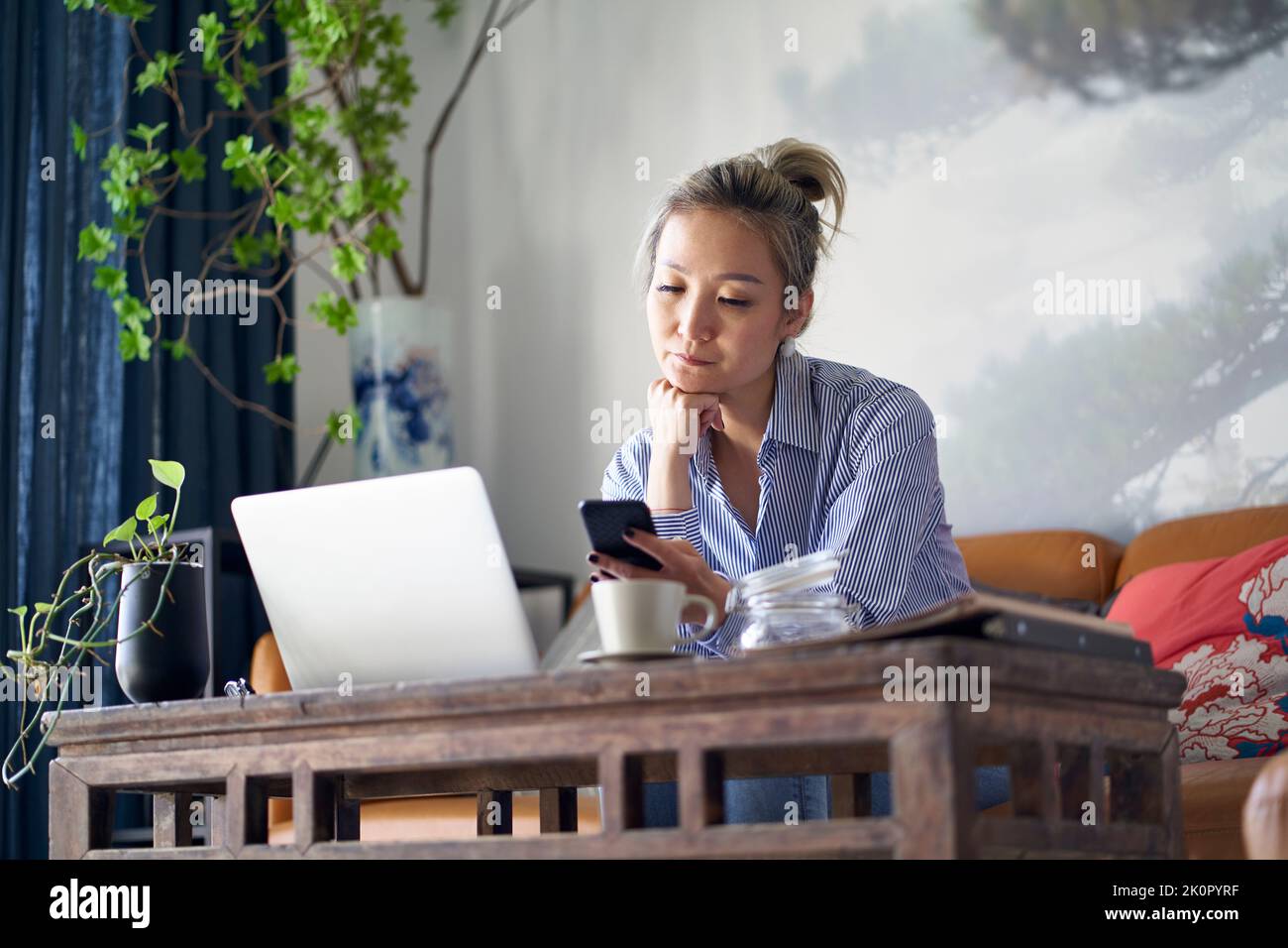 Reife professionelle asiatische Frau sitzt auf der Couch arbeiten von zu Hause aus mit Handy und Notebook-Computer Stockfoto