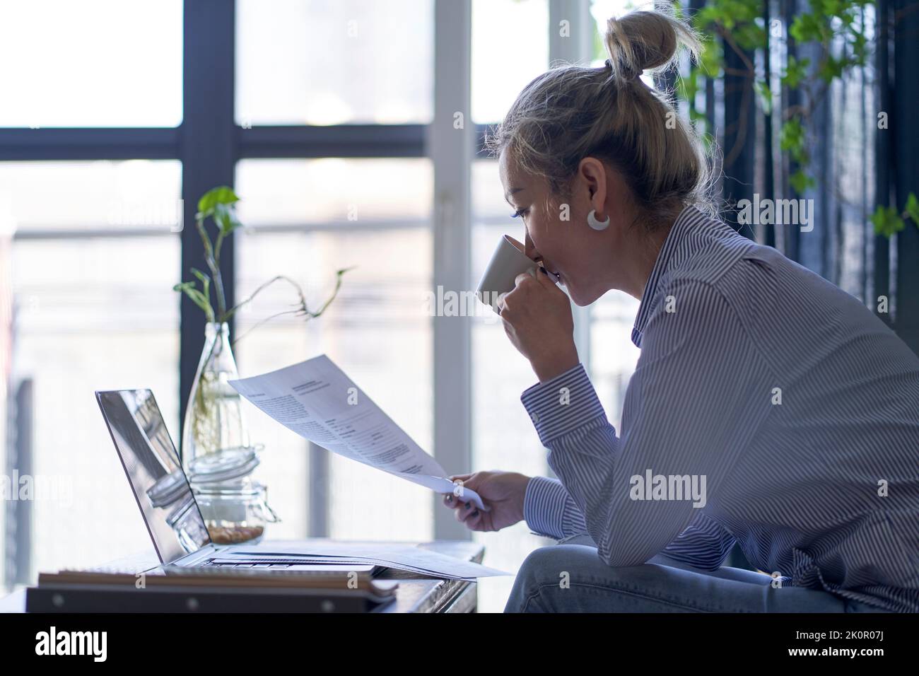 Reife professionelle asiatische Frau, die von zu Hause aus mit einem Laptop-Computer arbeitet, während sie Papierdokumente, Seitenansicht, ansieht Stockfoto